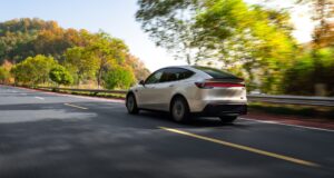 Tesla Model Y Long Wheelbase driving on a tree-lined road, rear three-quarter view