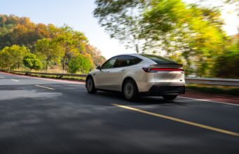 Tesla Model Y Long Wheelbase Brings Six Seats to the US for $61,990 Tesla Model Y Long Wheelbase driving on a tree-lined road, rear three-quarter view