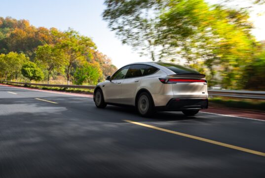 Tesla Model Y Long Wheelbase driving on a tree-lined road, rear three-quarter view