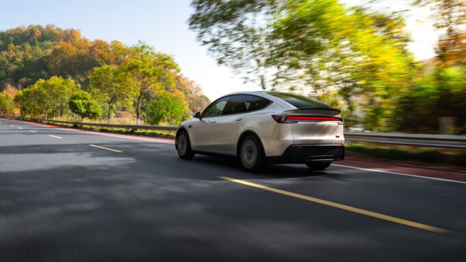 Tesla Model Y Long Wheelbase Brings Six Seats to the US for $61,990 Tesla Model Y Long Wheelbase driving on a tree-lined road, rear three-quarter view