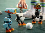 Pollen Robotics Microduck Opens Preorders at 399 Dollars Three Microduck robots on a turf pitch around a small ball, with a hand steadying one unit