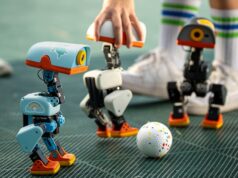 Pollen Robotics Microduck Opens Preorders at 399 Dollars Three Microduck robots on a turf pitch around a small ball, with a hand steadying one unit