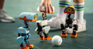 Three Microduck robots on a turf pitch around a small ball, with a hand steadying one unit