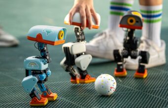 Pollen Robotics Microduck Opens Preorders at 399 Dollars Three Microduck robots on a turf pitch around a small ball, with a hand steadying one unit