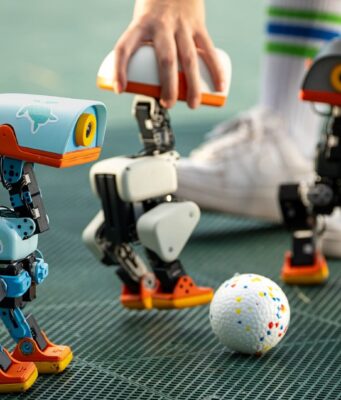 Pollen Robotics Microduck Opens Preorders at 399 Dollars Three Microduck robots on a turf pitch around a small ball, with a hand steadying one unit