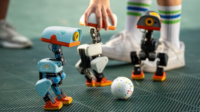 featured-pollen-microduck-kickabout Three Microduck robots on a turf pitch around a small ball, with a hand steadying one unit