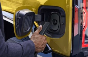 Every 2027 GM EV Will Plug Into Tesla Superchargers Official GM photo of a NACS charger plug at the charge port of a yellow GMC Hummer EV