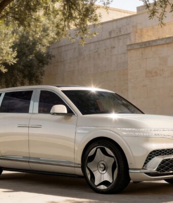 Genesis GV90 Neolun Flagship EV SUV Debuts With Coach Doors Cream Genesis GV90 three-quarter front view beside a stone wall and olive tree