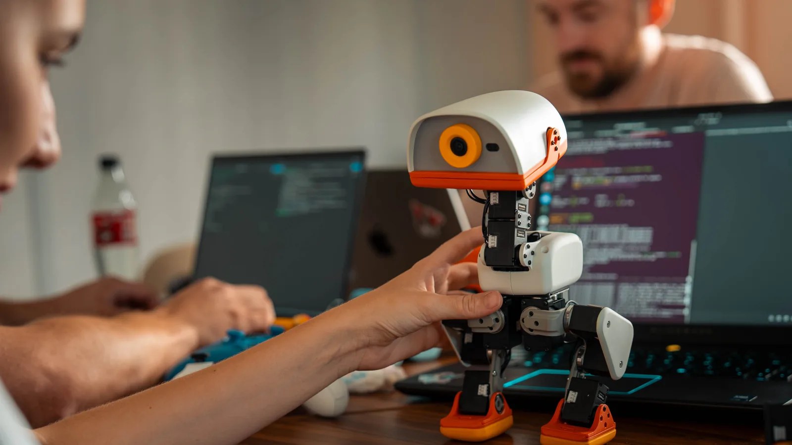 A hand holding a cream Microduck upright on a desk between two laptops