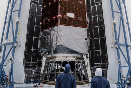 Technicians in blue clean-room suits stand before NASA's Nancy Grace Roman Space Telescope inside open Falcon Heavy fairing halves
