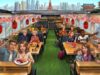 Illustrated daytime cabin of the Sushi Bus with a sushi conveyor, lanterns, and a Tokyo skyline