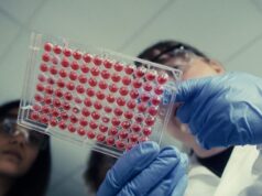 Anthropic Wet Lab Confirmed: Claude Maker Now Runs Physical Biology Experiments Two scientists in safety glasses and blue gloves hold up a 96-well plate filled with red liquid in a lab
