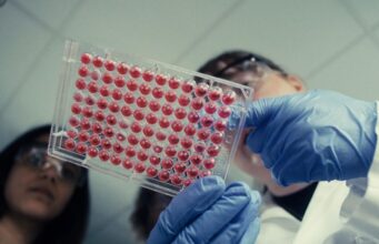 Anthropic Wet Lab Confirmed: Claude Maker Now Runs Physical Biology Experiments Two scientists in safety glasses and blue gloves hold up a 96-well plate filled with red liquid in a lab