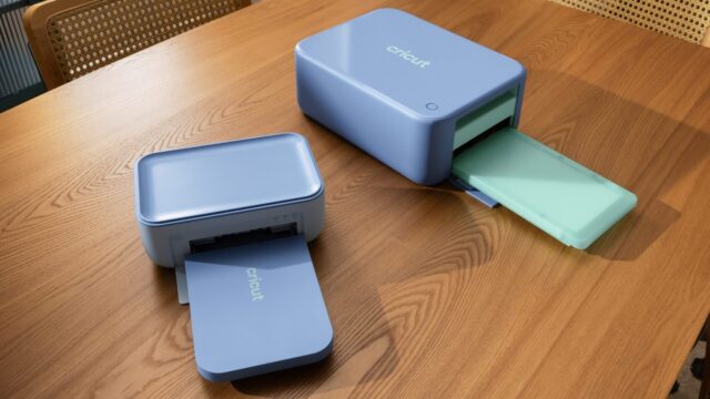 Cricut StickerPix Print and Print + Cut machines on a wooden table with output trays open