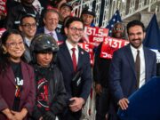 Mayor Zohran Mamdani and officials standing with delivery workers holding red $131.5 million signs at City Hall