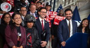Mayor Zohran Mamdani and officials standing with delivery workers holding red $131.5 million signs at City Hall