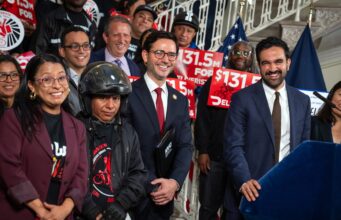 Mayor Zohran Mamdani and officials standing with delivery workers holding red $131.5 million signs at City Hall