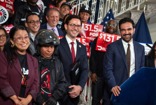 Mayor Zohran Mamdani and officials standing with delivery workers holding red $131.5 million signs at City Hall