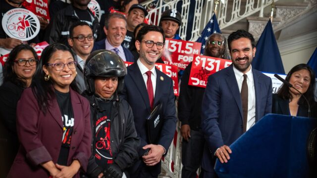 doordash-nyc-settlement-city-hall Mayor Zohran Mamdani and officials standing with delivery workers holding red $131.5 million signs at City Hall