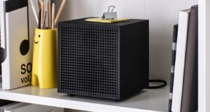 Black IKEA Badkruka cube speaker on a white shelf between books and a yellow pencil holder, with its power cable behind it