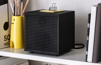IKEA Badkruka Speaker: $59.99 Wired Bluetooth Audio Black IKEA Badkruka cube speaker on a white shelf between books and a yellow pencil holder, with its power cable behind it