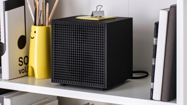 Black IKEA Badkruka cube speaker on a white shelf between books and a yellow pencil holder, with its power cable behind it