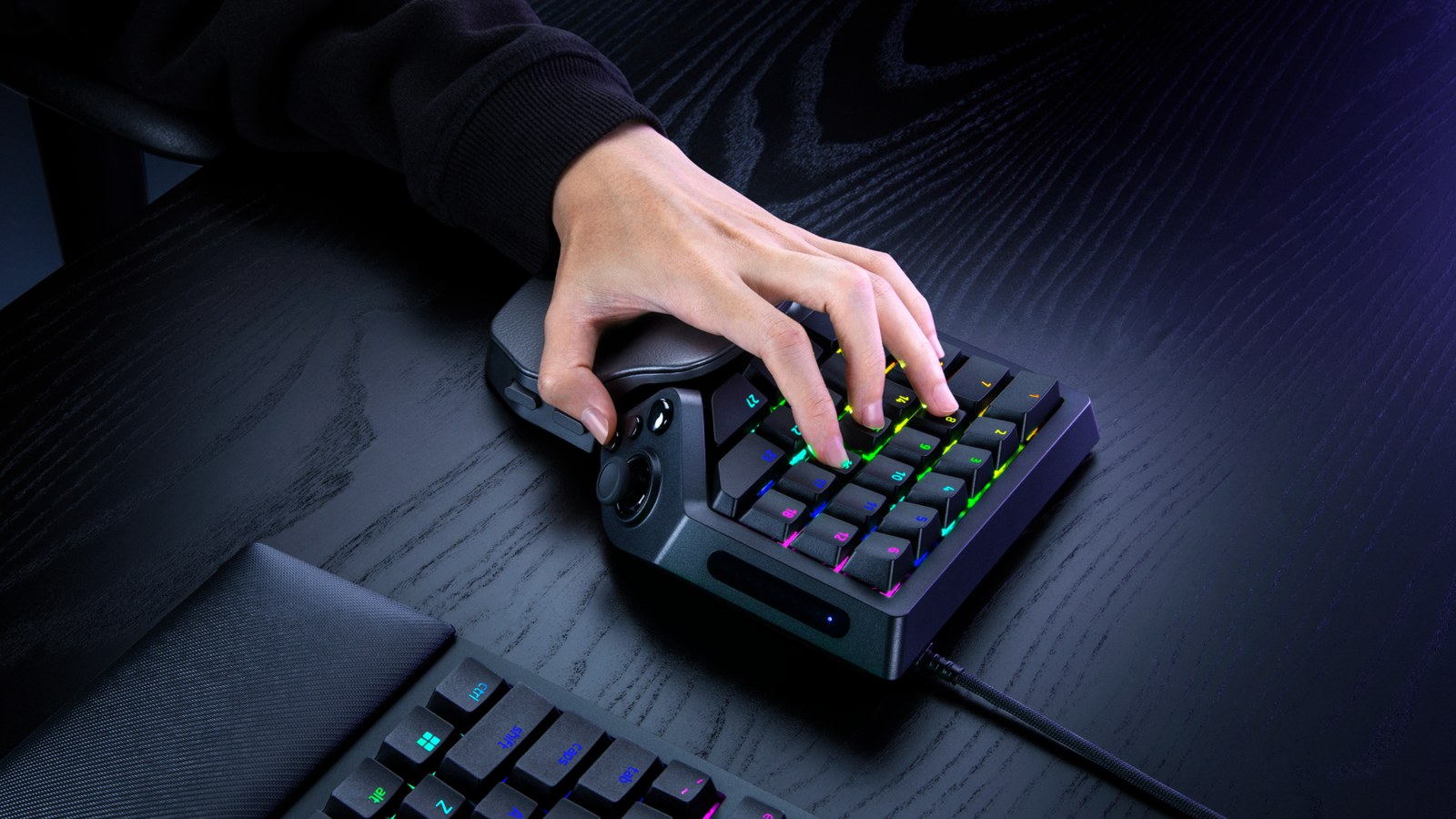 A hand resting on the Razer Tartarus V2 Pro keypad with RGB-lit numbered keys, next to a Razer keyboard on a dark desk