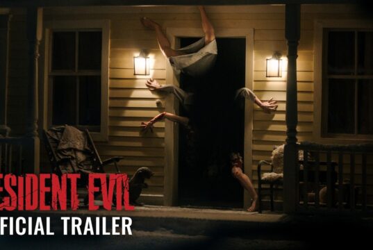 A figure contorted upside down in the doorway of a lit farmhouse porch at night, from Sony's Resident Evil official trailer