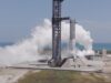 A SpaceX Super Heavy booster on the launch mount beside the launch tower at Starbase, wrapped in white vapor with the Gulf behind it