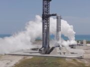 A SpaceX Super Heavy booster on the launch mount beside the launch tower at Starbase, wrapped in white vapor with the Gulf behind it