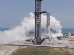 A SpaceX Super Heavy booster on the launch mount beside the launch tower at Starbase, wrapped in white vapor with the Gulf behind it