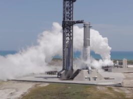 A SpaceX Super Heavy booster on the launch mount beside the launch tower at Starbase, wrapped in white vapor with the Gulf behind it