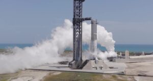 A SpaceX Super Heavy booster on the launch mount beside the launch tower at Starbase, wrapped in white vapor with the Gulf behind it