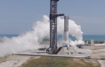 A SpaceX Super Heavy booster on the launch mount beside the launch tower at Starbase, wrapped in white vapor with the Gulf behind it