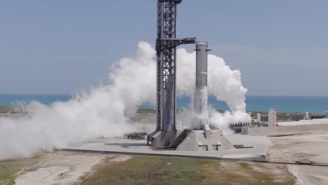 A SpaceX Super Heavy booster on the launch mount beside the launch tower at Starbase, wrapped in white vapor with the Gulf behind it