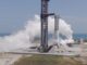 A SpaceX Super Heavy booster on the launch mount beside the launch tower at Starbase, wrapped in white vapor with the Gulf behind it