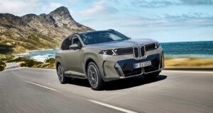 2027 BMW iX5 60 xDrive driving along a coastal mountain road showing its illuminated vertical kidney grille and B-pillar Winglet door handles