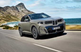 2027 BMW iX5 60 xDrive Starts at $79,800 With an Estimated 435-Mile Range and 460 kW Charging 2027 BMW iX5 60 xDrive driving along a coastal mountain road showing its illuminated vertical kidney grille and B-pillar Winglet door handles