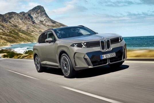 2027 BMW iX5 60 xDrive driving along a coastal mountain road showing its illuminated vertical kidney grille and B-pillar Winglet door handles