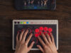 8BitDo Arcade Controller Pro on a dark wooden desk, a player's hands resting on its round red and black buttons, with a Nintendo Switch console showing a fighting game at the top of the frame