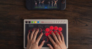 8BitDo Arcade Controller Pro on a dark wooden desk, a player's hands resting on its round red and black buttons, with a Nintendo Switch console showing a fighting game at the top of the frame