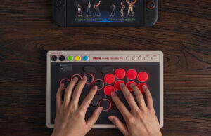 8BitDo Arcade Controller Pro on a dark wooden desk, a player's hands resting on its round red and black buttons, with a Nintendo Switch console showing a fighting game at the top of the frame