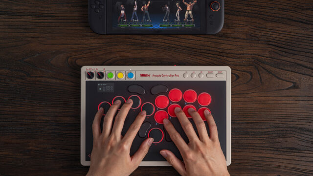 8BitDo Arcade Controller Pro on a dark wooden desk, a player's hands resting on its round red and black buttons, with a Nintendo Switch console showing a fighting game at the top of the frame