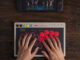 8BitDo Arcade Controller Pro US Pre-Order Opens at $119.99 8BitDo Arcade Controller Pro on a dark wooden desk, a player's hands resting on its round red and black buttons, with a Nintendo Switch console showing a fighting game at the top of the frame