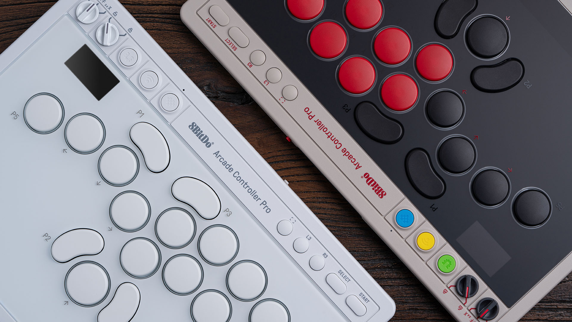 Two 8BitDo Arcade Controller Pro pads side by side on a wooden desk, one with white buttons on a white chassis and one with red and black buttons on a cream chassis, each showing a small display and control-panel dials
