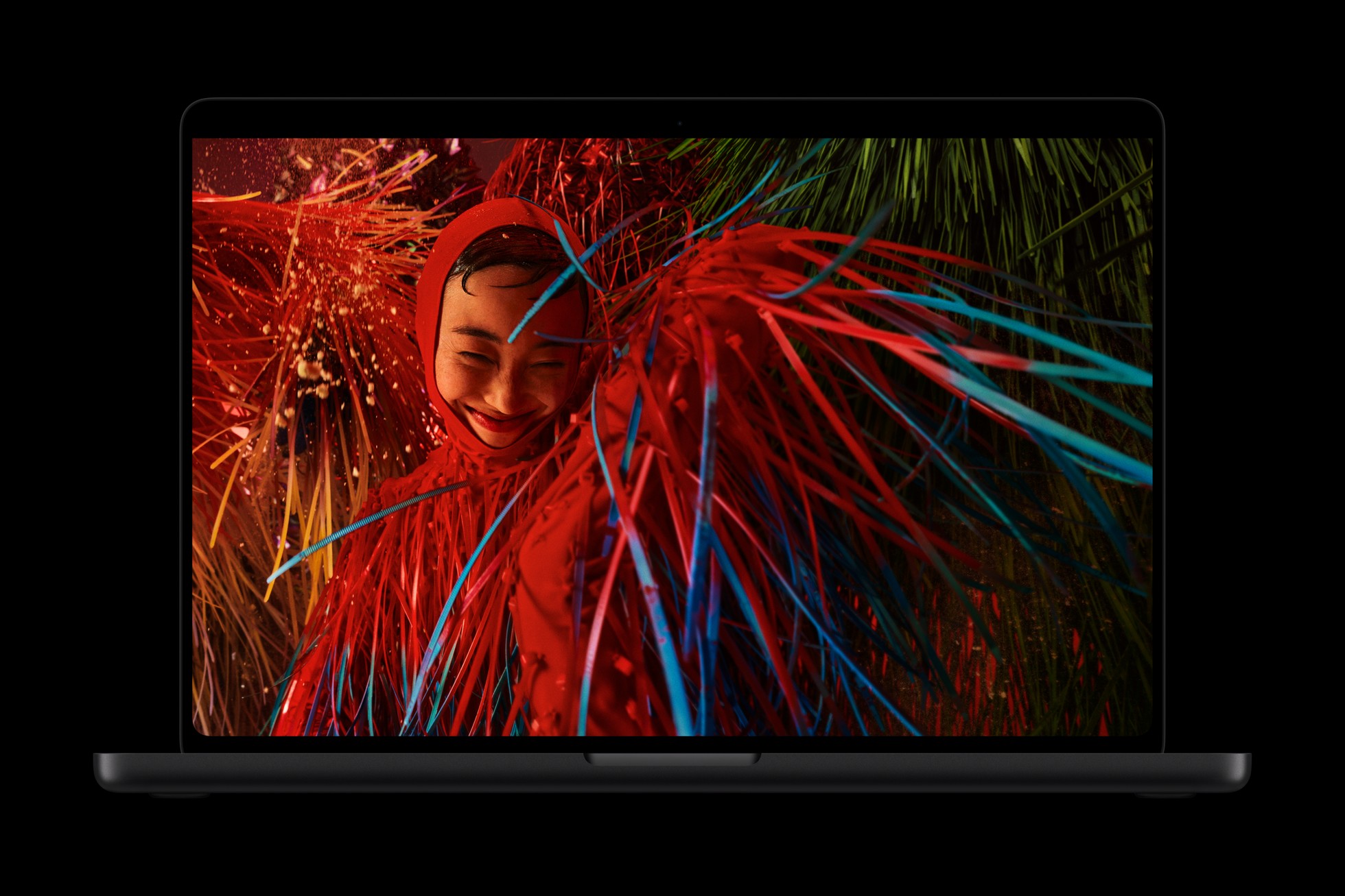 An open MacBook Pro with PROLED XDR Display shows a saturated photo of a smiling person in a red hood surrounded by red and cyan filaments