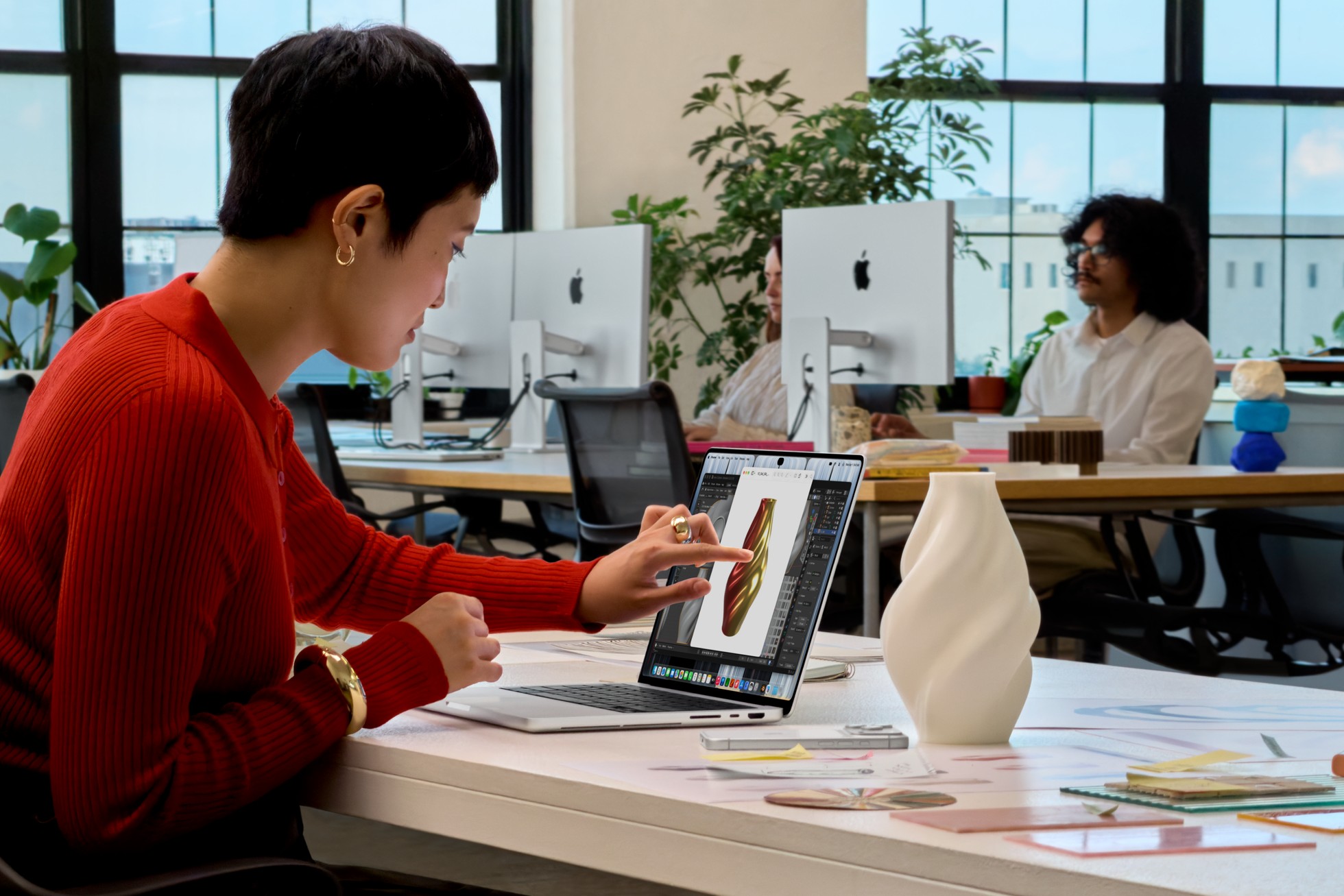 A person in a red top touches the screen of a MacBook Pro with PROLED XDR Display showing a 3D modelling app in an open-plan office