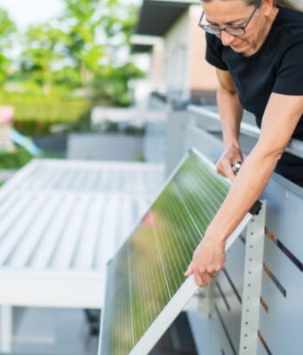California Plug-In Solar Law Skips Utility Approval, but Caps Systems at 1,200 Watts Woman attaching a plug-in solar panel to the outside of a balcony railing