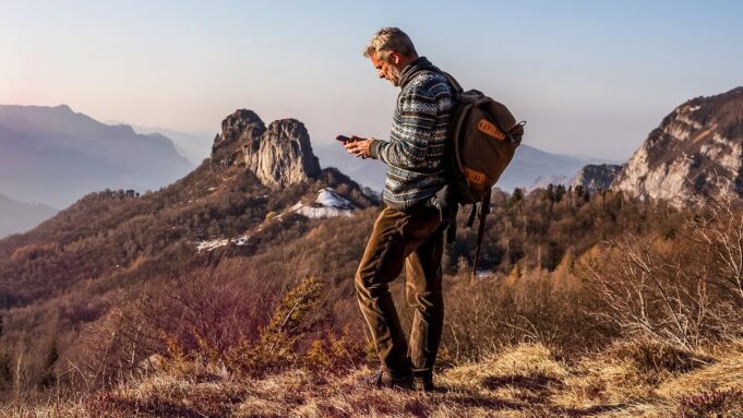 Satellite Joint Venture From AT&T, T-Mobile and Verizon Has a CEO but No Launch Date Hiker with a backpack using a smartphone on a remote mountain ridge at sunset