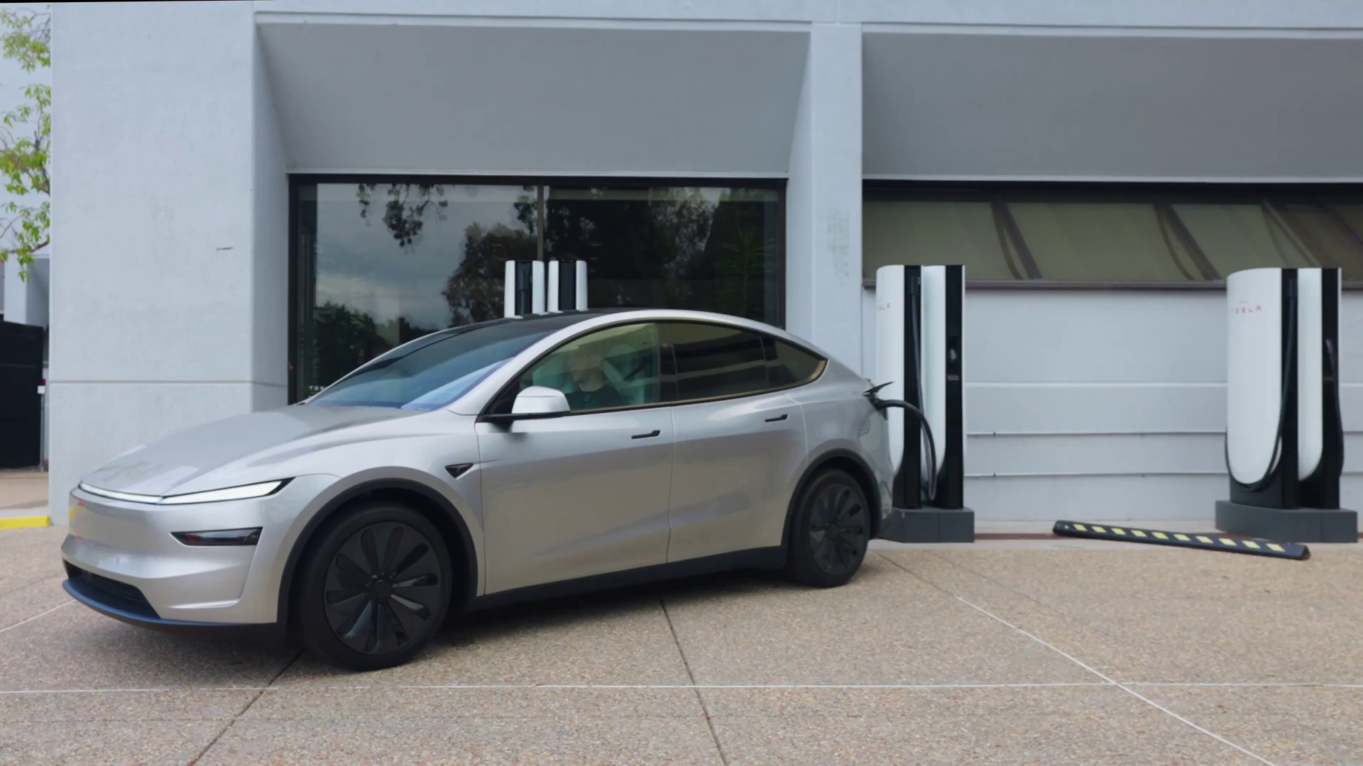 Silver Tesla Model Y driving away from a Supercharger stall with the charging cable still attached